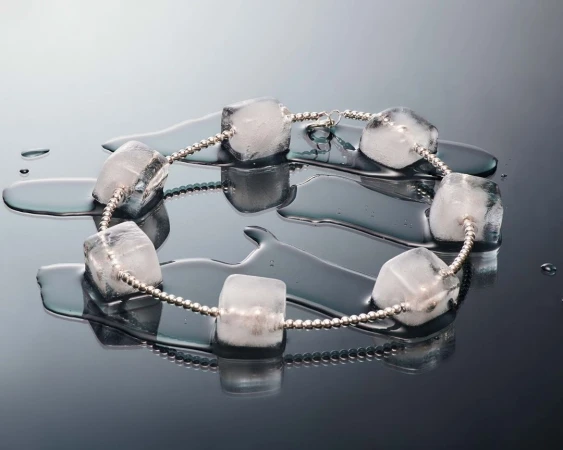 Still life of clear ice cube beads on a silver beaded string, reflected on a black surface, showing a pattern of cubes and water droplets.