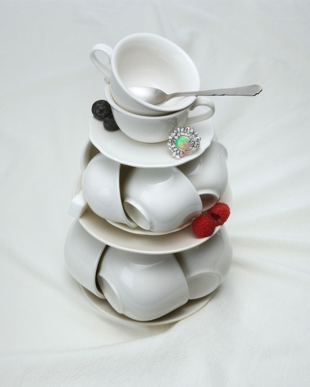 Stacked white teacups and saucers tower adorned with a spoon, berries, and a ring, creating a whimsical still life on a white fabric background.