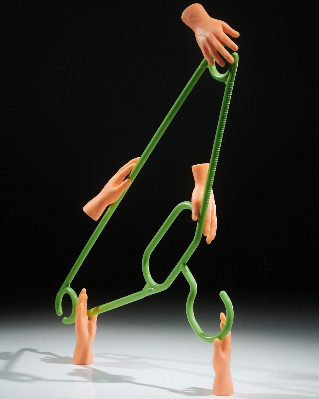 A green clothes hanger is held by six disembodied mannequin hands against a dark backdrop, creating a surreal and intriguing composition.