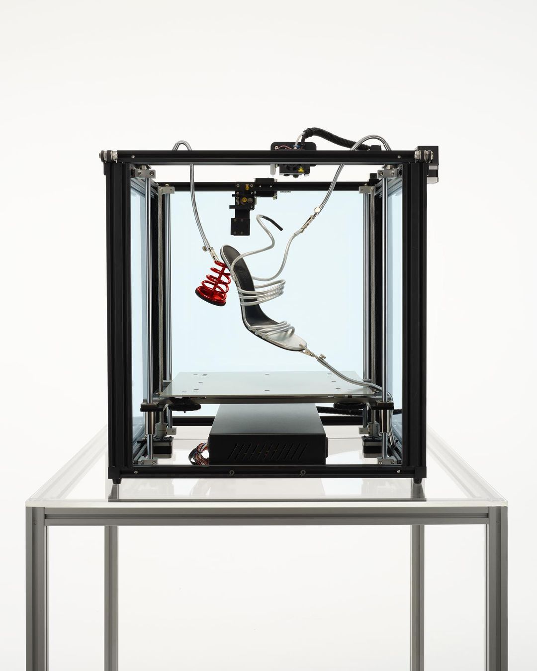 Silver high heel shoe positioned inside a black metal frame with a red spring attached and resting on a reflective surface.