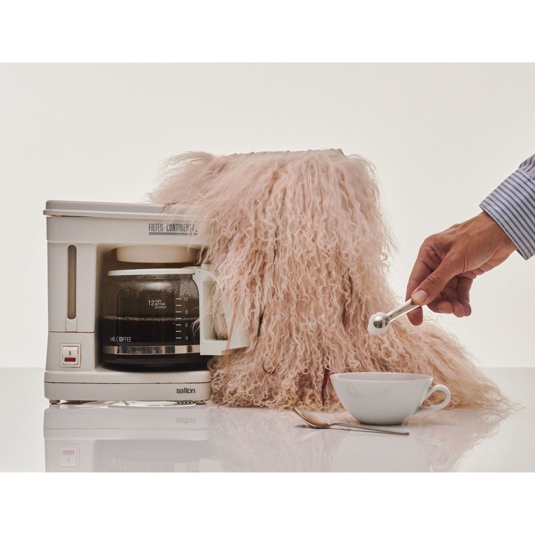 Coffee maker topped with furry pink throw with a hand holding a silver scoop above a white cup with a spoon on a white table.