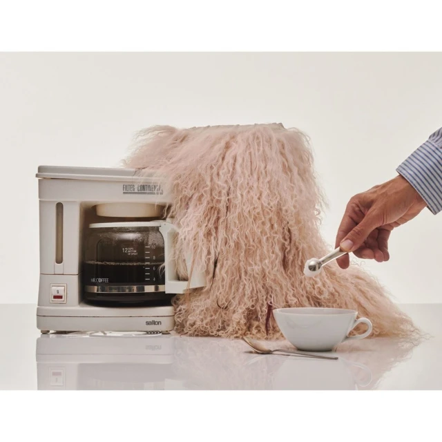 Coffee maker topped with furry pink throw with a hand holding a silver scoop above a white cup with a spoon on a white table.