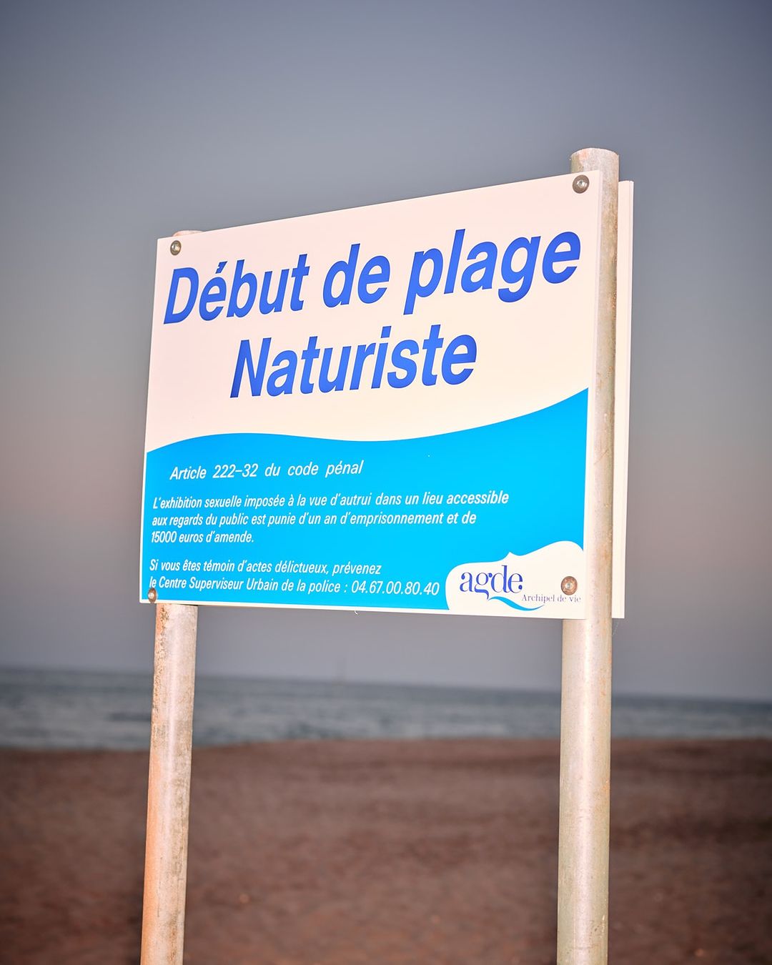 A sign that indicates the beginning of a naturist beach, with white sand and ocean water in the background on a cloudy day.