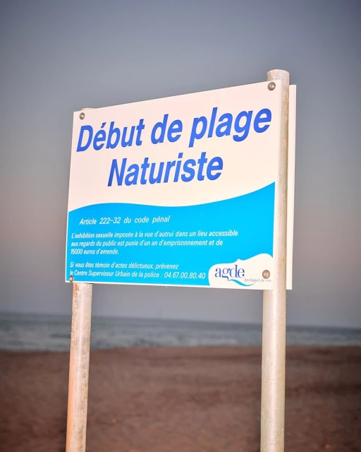 A sign that indicates the beginning of a naturist beach, with white sand and ocean water in the background on a cloudy day.