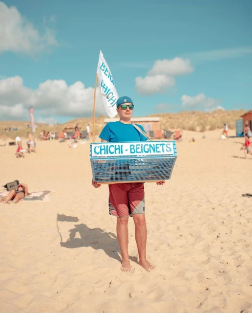 Beignet vendor in a blue shirt, hat, and sunglasses standing on a sandy beach carrying a blue container with 'Chichi - Beignets' sign.