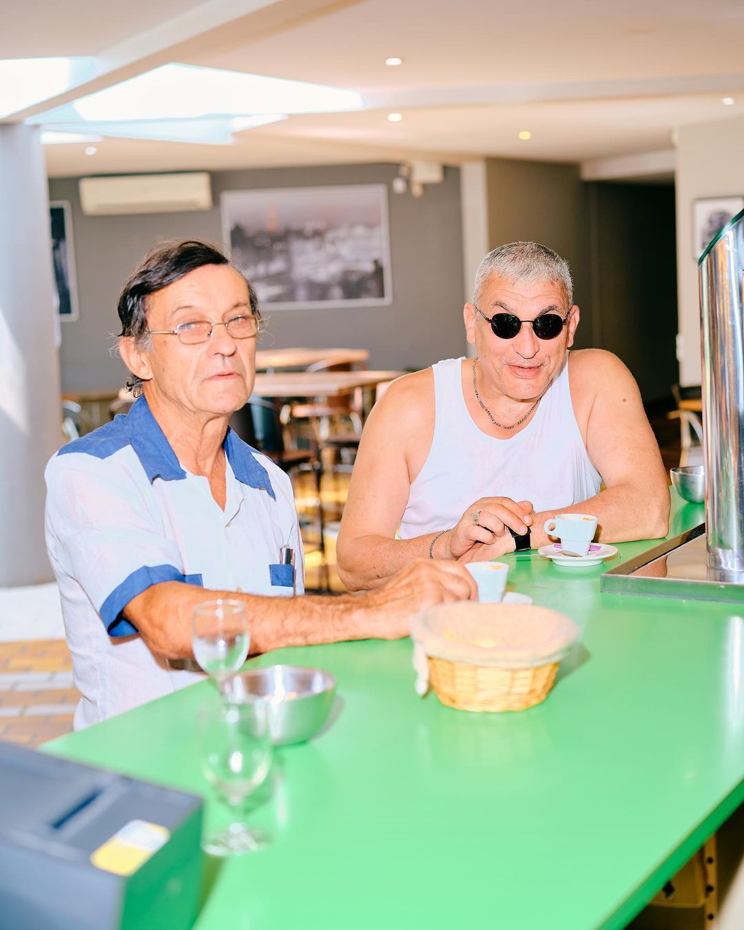 Two older men sit at a green counter in an indoor setting. One wears sunglasses. Coffee cups and silverware are on the counter.
