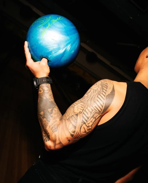 Man holding a blue bowling ball with tattoos on his arm in a dimly lit indoor setting, focusing on sports and body art.