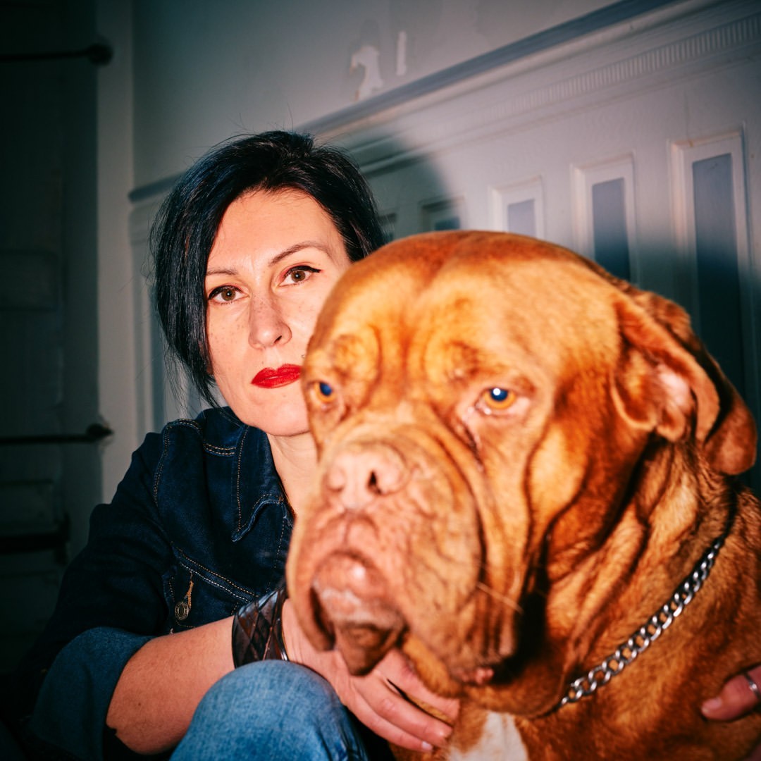 A woman with short dark hair embraces her dogue de bordeaux in an indoor setting with a soft color palette and close-up framing.