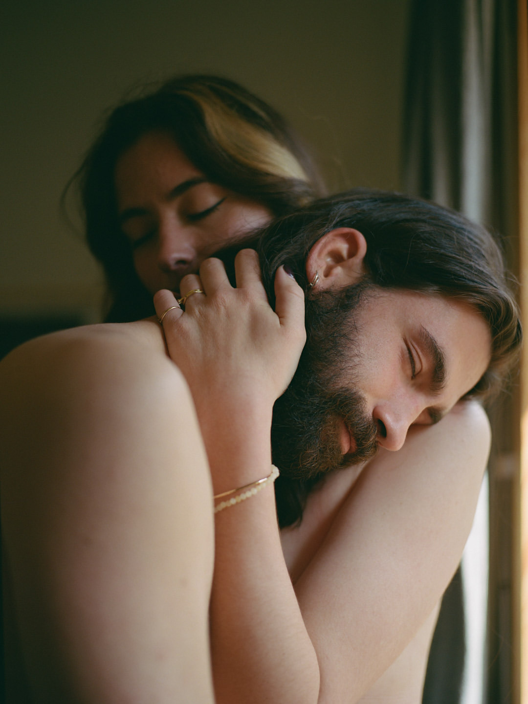 A woman embraces a man with his head on her shoulder indoors, both with eyes closed in a close-up studio portrait.