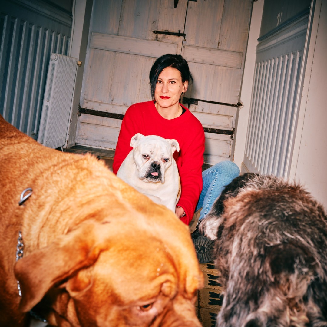 Woman in red sweater sits indoors with three dogs of different breeds, one tan, one white, and one with grey and brown fur.