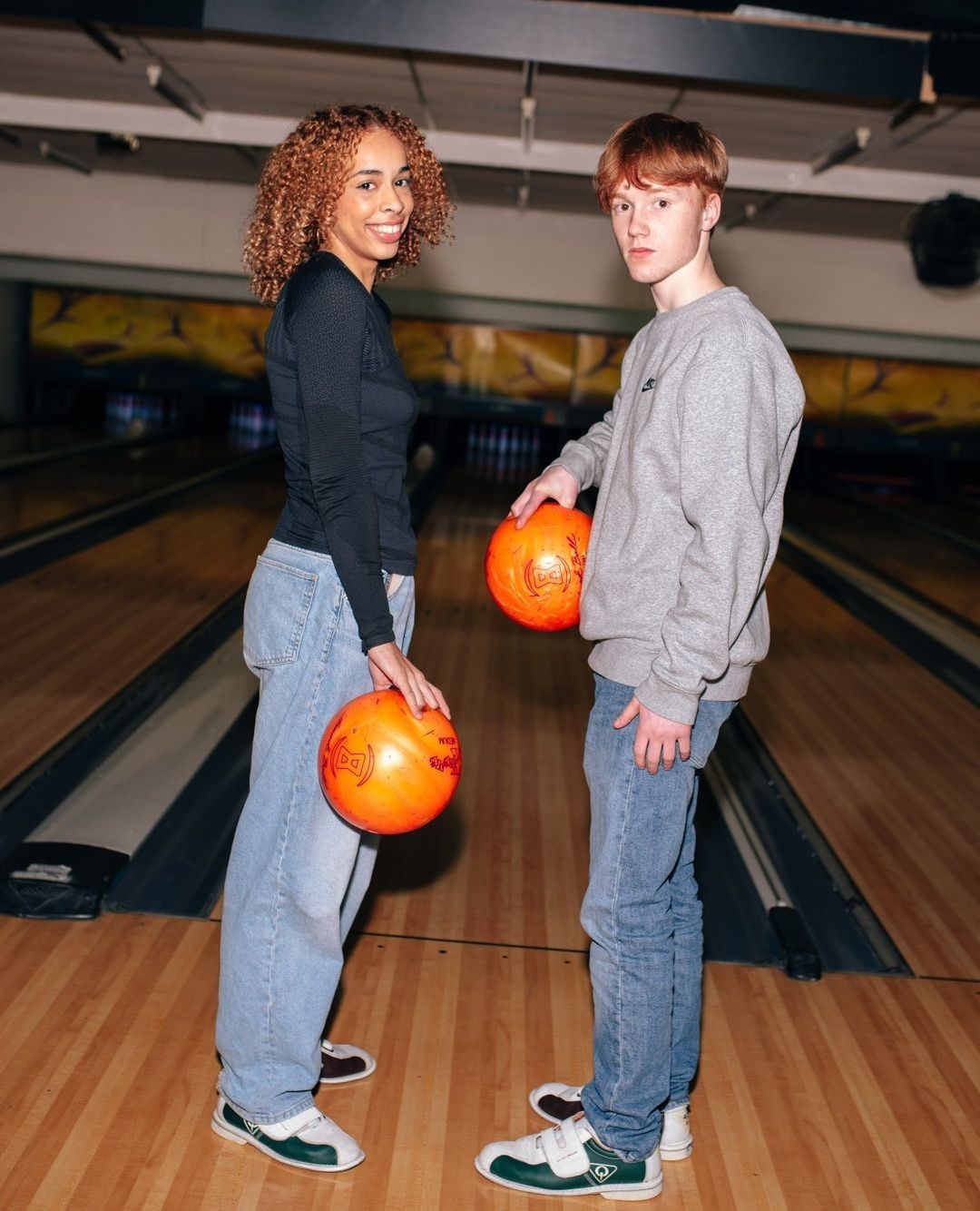 Two smiling people in bowling alley holding orange bowling balls. The bowling alley background is visible with bowling lanes.