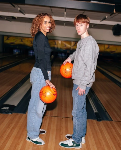 Two smiling people in bowling alley holding orange bowling balls. The bowling alley background is visible with bowling lanes.
