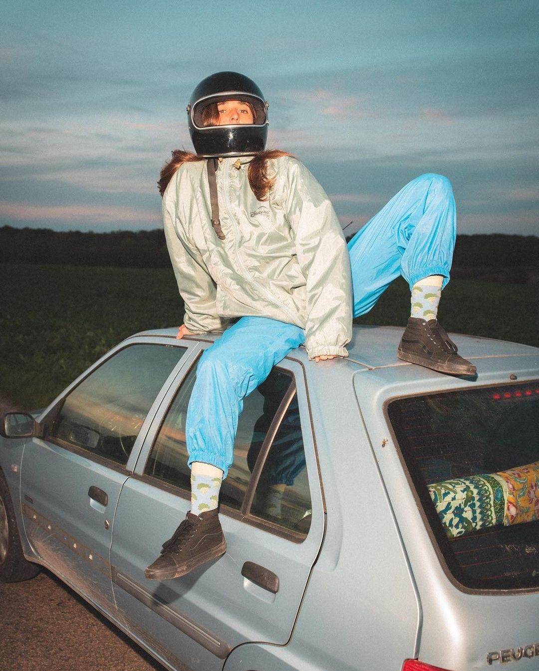 Person wearing a helmet and jacket sitting on a silver car against a twilight sky and a green field in an outdoor setting.