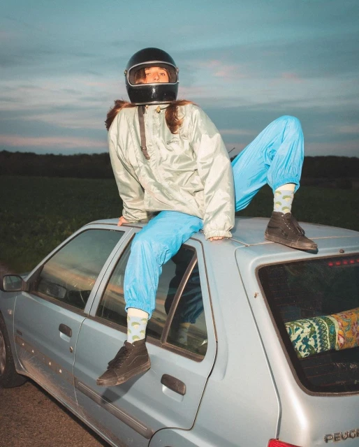 Person wearing a helmet and jacket sitting on a silver car against a twilight sky and a green field in an outdoor setting.