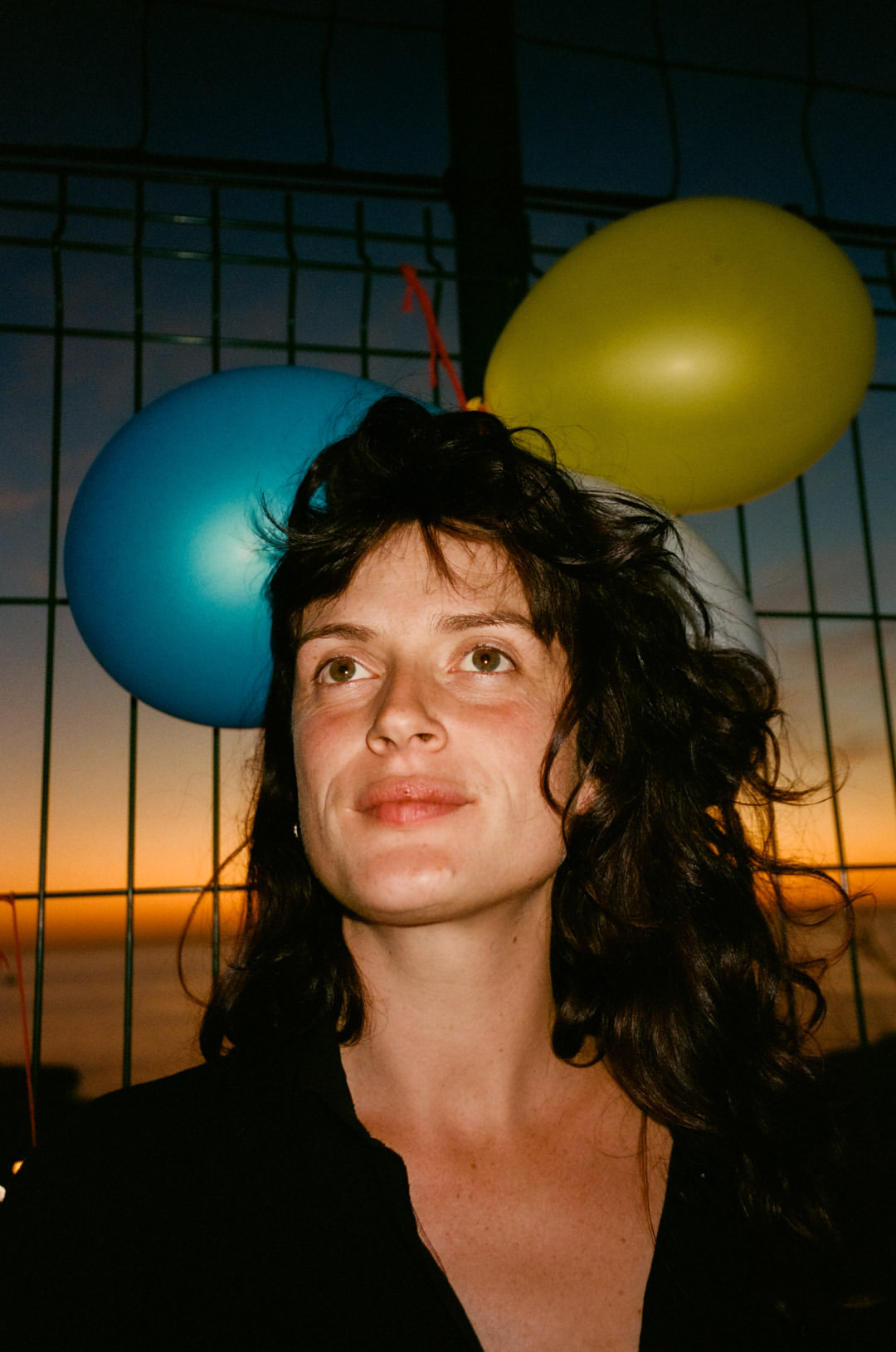 Portrait of a woman with dark hair and balloons, against a fence with a sunset and ocean backdrop at dusk. Close up.