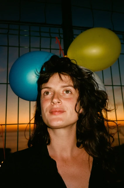 Portrait of a woman with dark hair and balloons, against a fence with a sunset and ocean backdrop at dusk. Close up.