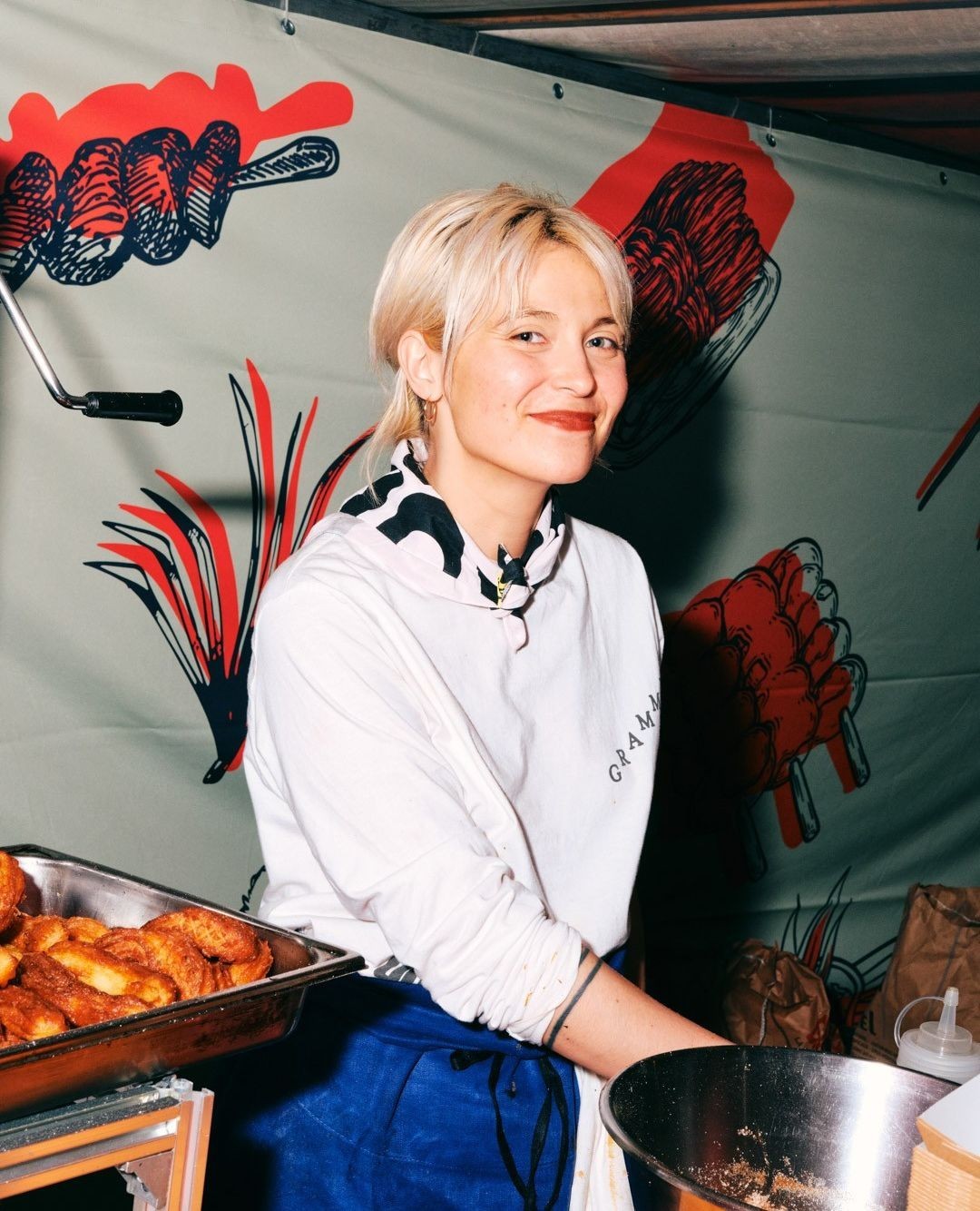 Smiling woman with short blonde hair stands at food stall with tray of pastries and graphic backdrop in a portrait style shot.