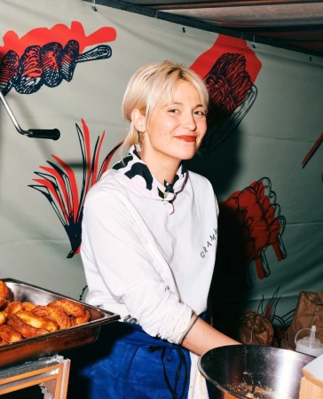 Smiling woman with short blonde hair stands at food stall with tray of pastries and graphic backdrop in a portrait style shot.