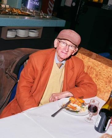 Elderly man in orange jacket and cap sits at a table with food and wine in a restaurant setting. Indoor dining with muted lighting.