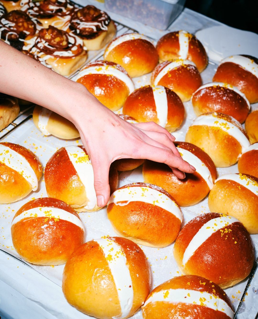 Overhead shot of hand selecting an iced hot cross bun from a tray with other buns and cinnamon rolls on a white surface.