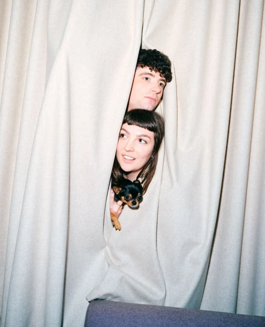 Two people peek through parted beige curtains while holding a small dog. The photo features an indoor setting with soft, diffused lighting.