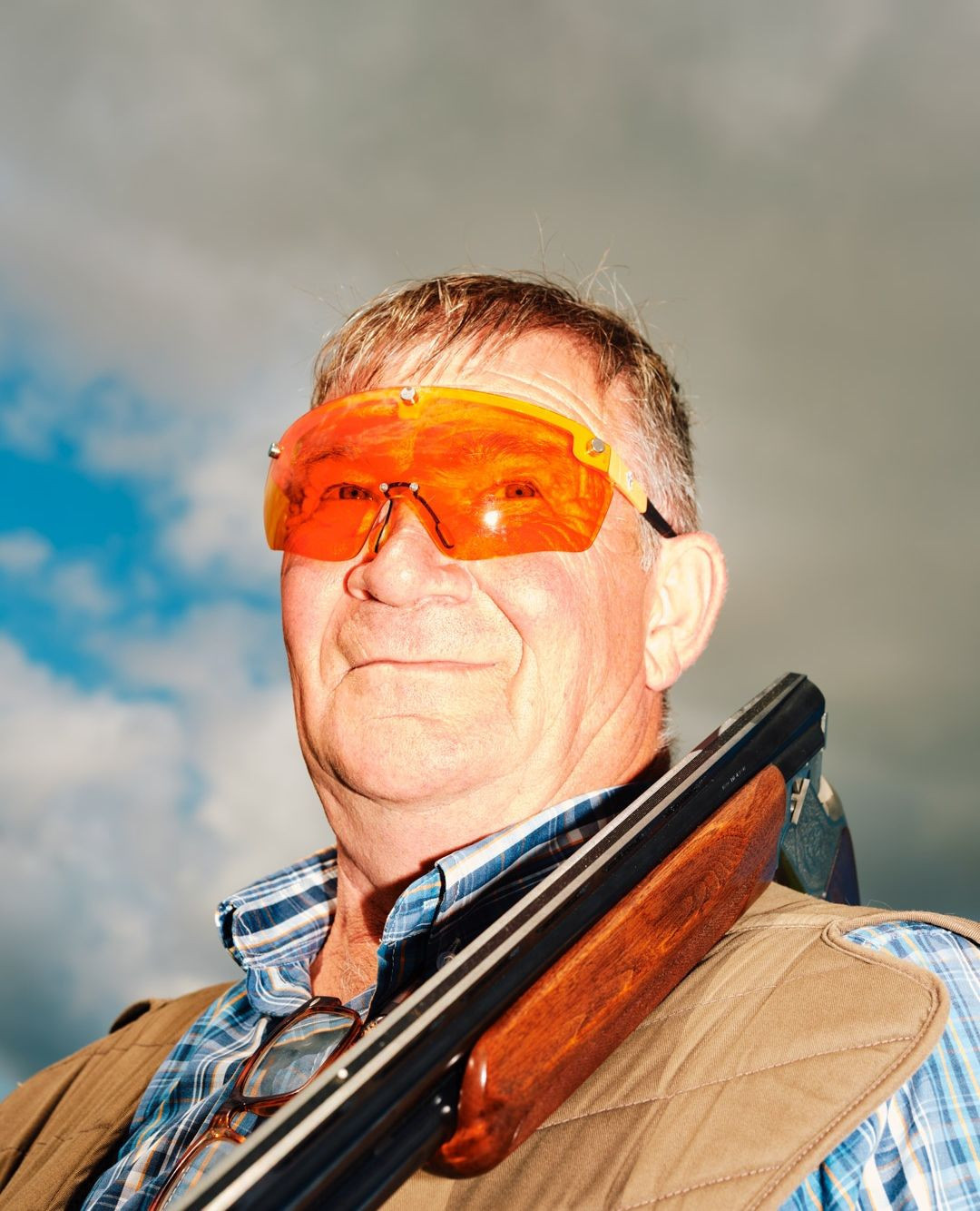 Older man outdoors with a shotgun wearing amber shooting glasses, a vest, and a plaid shirt, set against a cloudy sky.