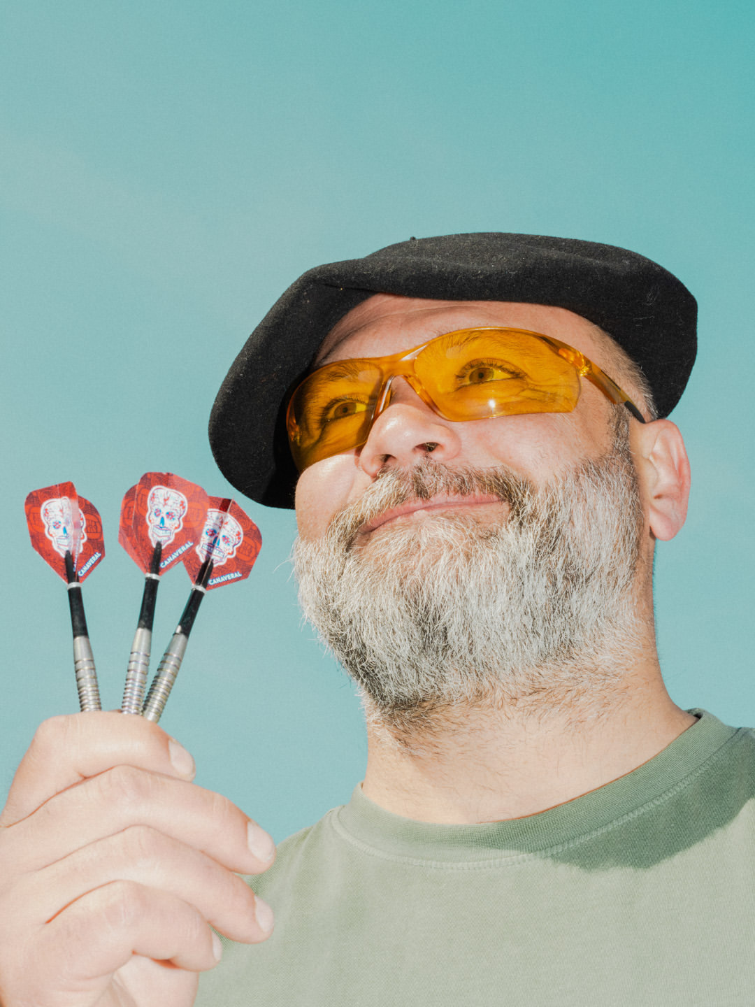 Adult male with a grey beard wearing a beret and yellow sunglasses holds three darts with skull designs against a turquoise backdrop.