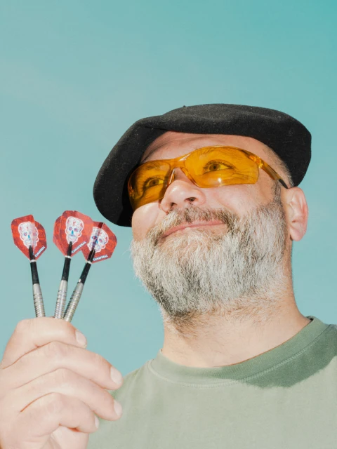 Adult male with a grey beard wearing a beret and yellow sunglasses holds three darts with skull designs against a turquoise backdrop.