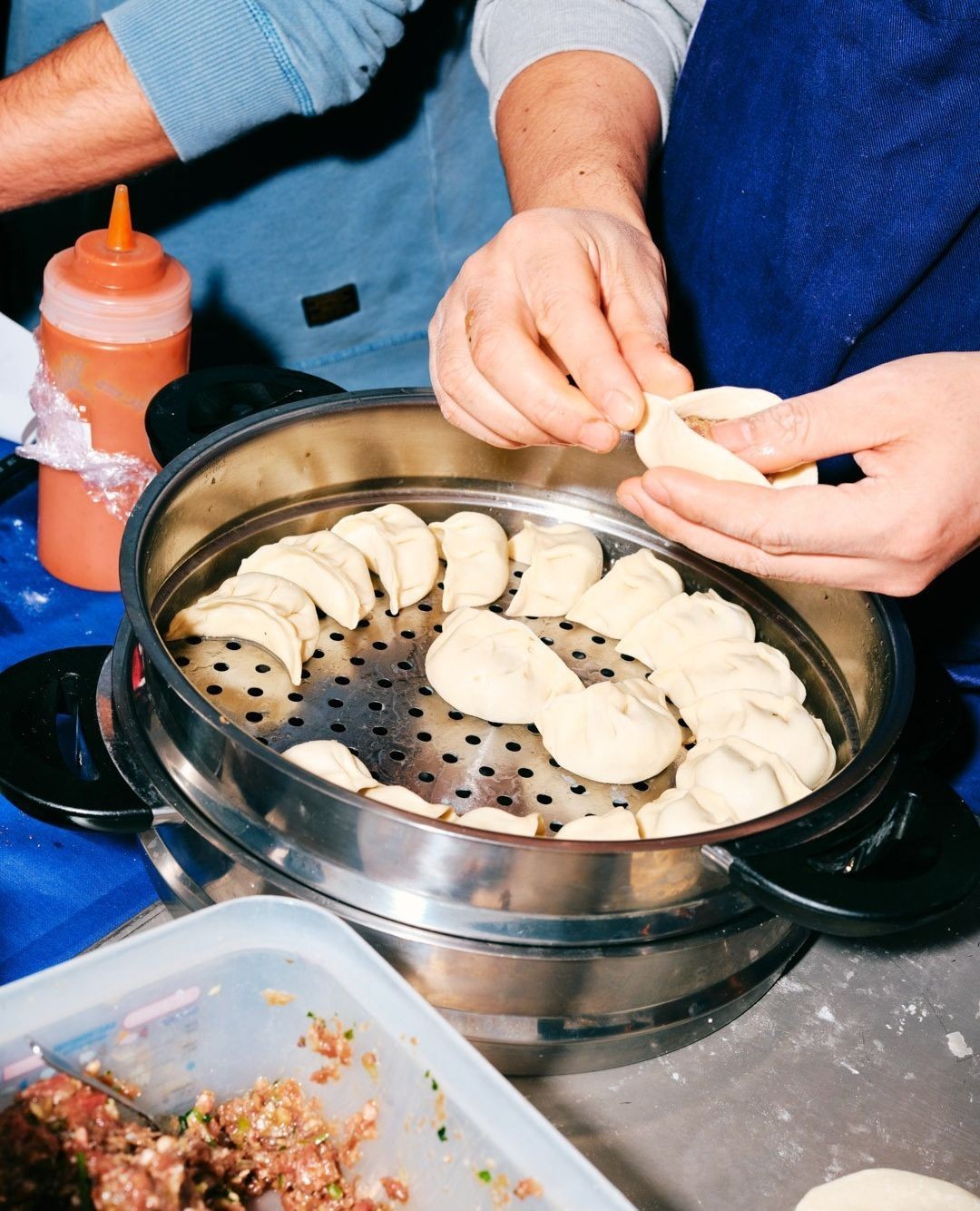Two adults preparing dumplings on a steel steamer on a table with other ingredients. Food and beverage photography.