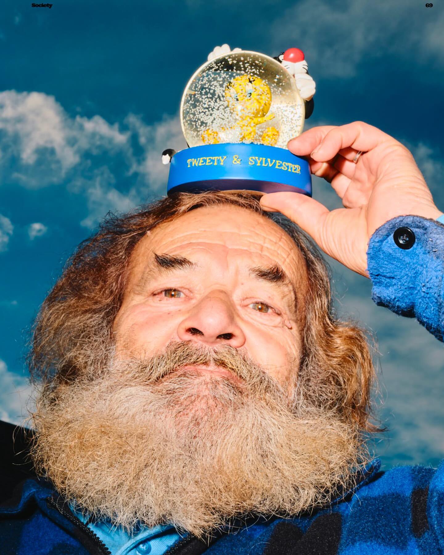 Portrait of an elderly man with a beard balancing a Tweety and Sylvester snow globe on his head against a cloudy blue sky backdrop.