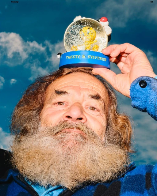 Portrait of an elderly man with a beard balancing a Tweety and Sylvester snow globe on his head against a cloudy blue sky backdrop.