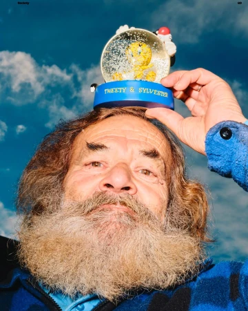 Portrait of an elderly man with a beard balancing a Tweety and Sylvester snow globe on his head against a cloudy blue sky backdrop.