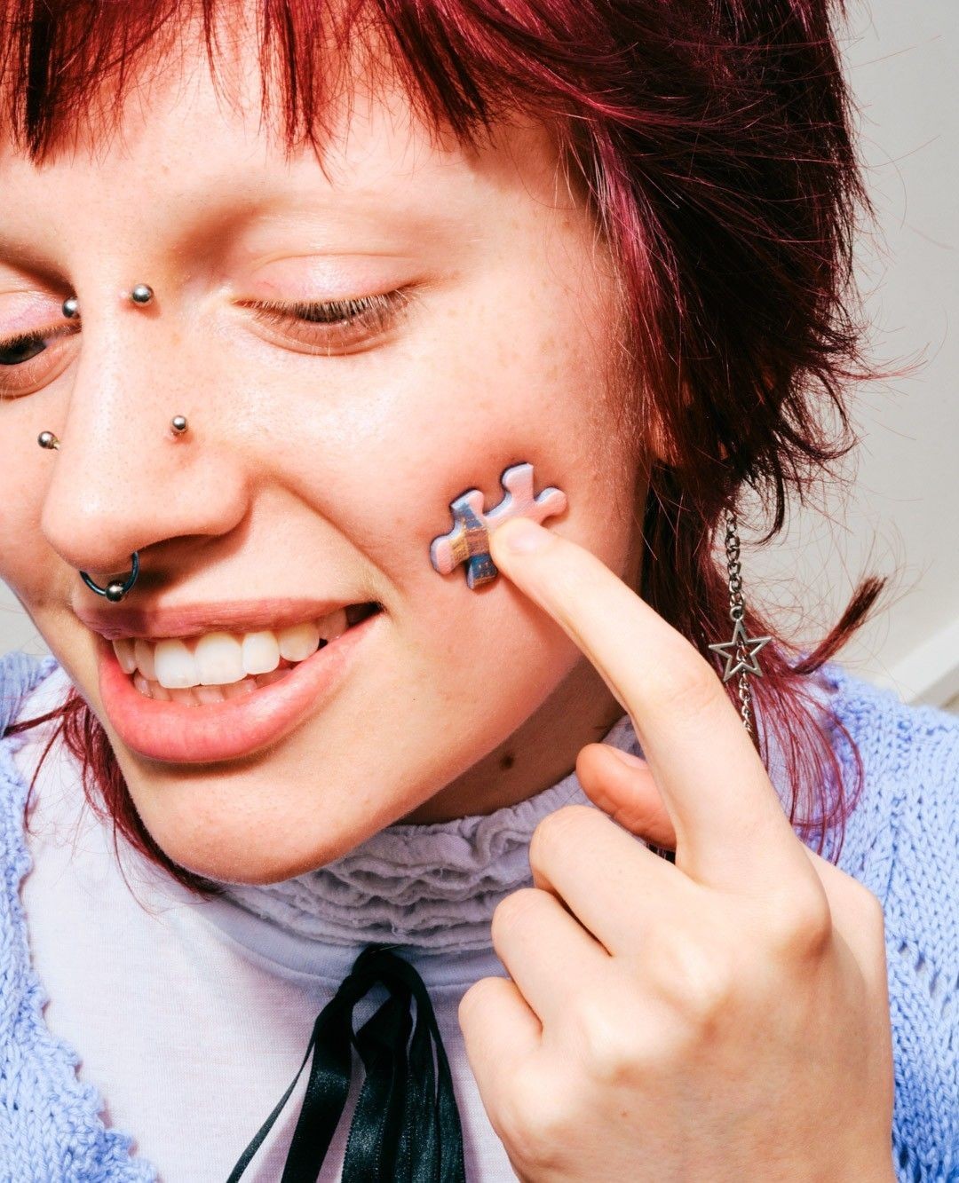 Close-up of a young adult woman with red hair holding a puzzle piece to her cheek, highlighting body modification and contemporary style.