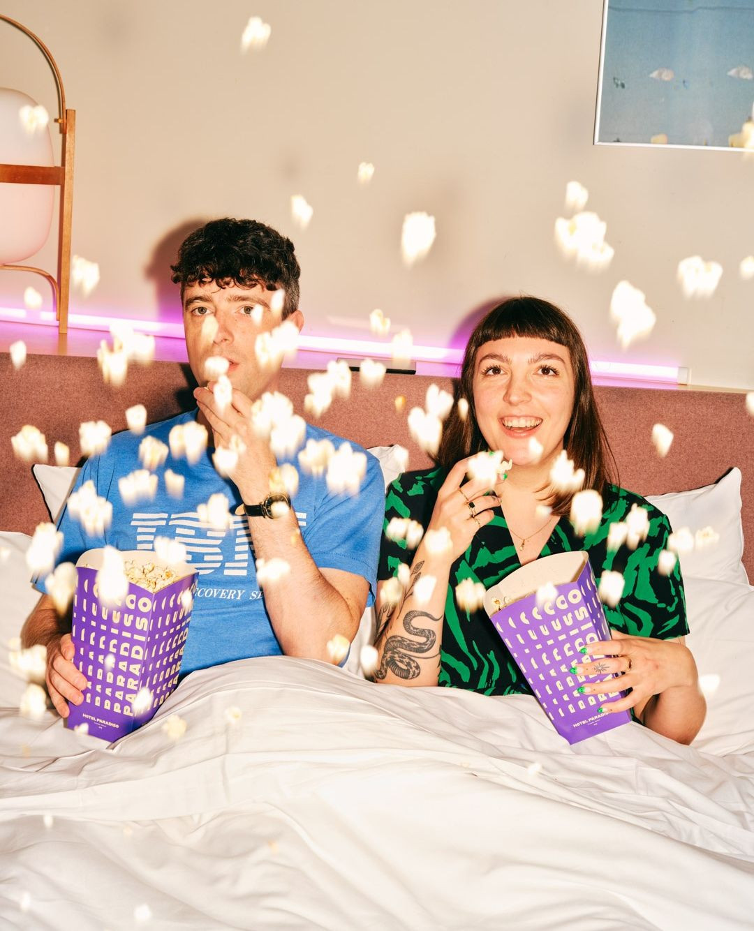 Young couple eating popcorn while lying in bed with white sheets and a pink headboard in a cozy indoor environment.