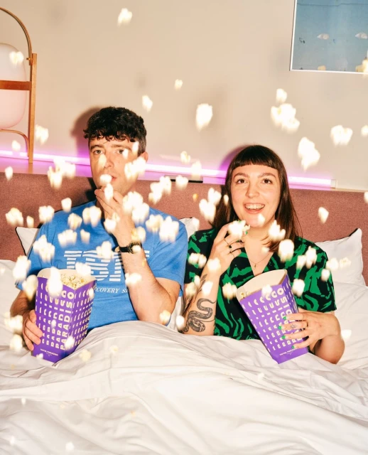 Young couple eating popcorn while lying in bed with white sheets and a pink headboard in a cozy indoor environment.