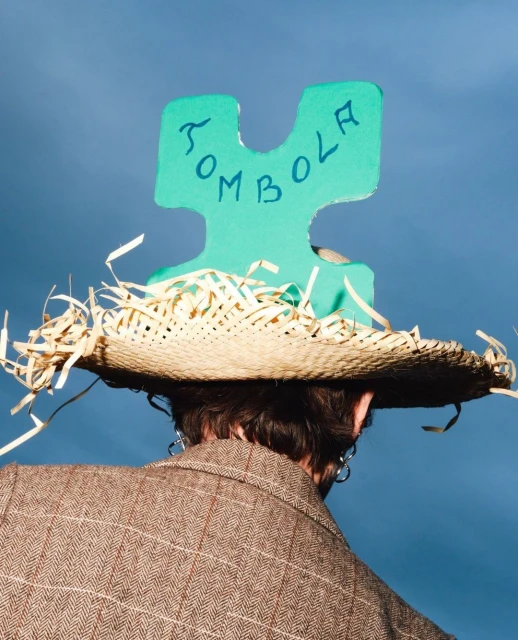 Person in a tweed jacket and straw hat, decorated with raffia and a green puzzle piece with TOMBOLA written, set against a blue sky.