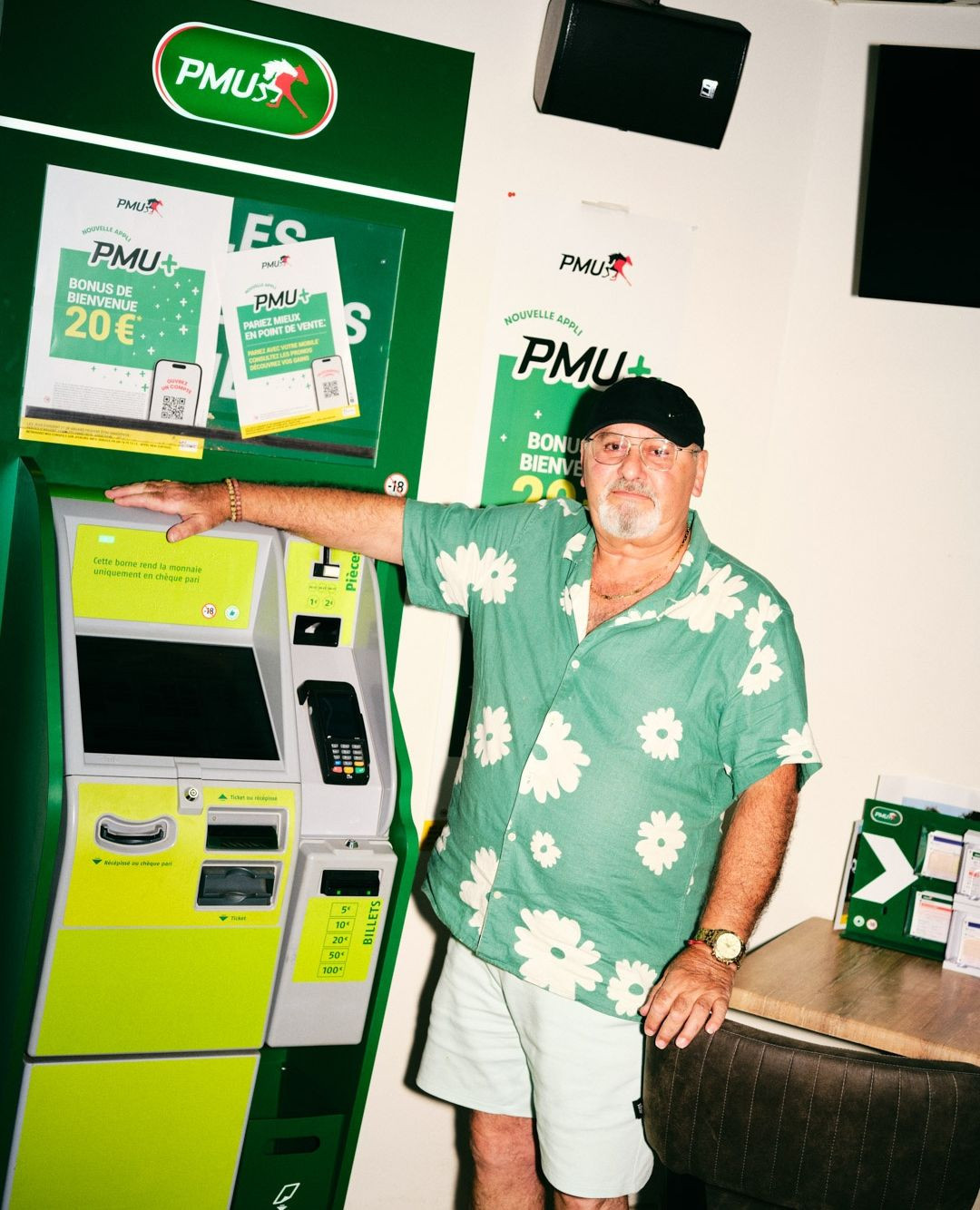 An adult man in a floral shirt stands beside a green and yellow PMU betting machine inside, with advertisements visible on the machine.