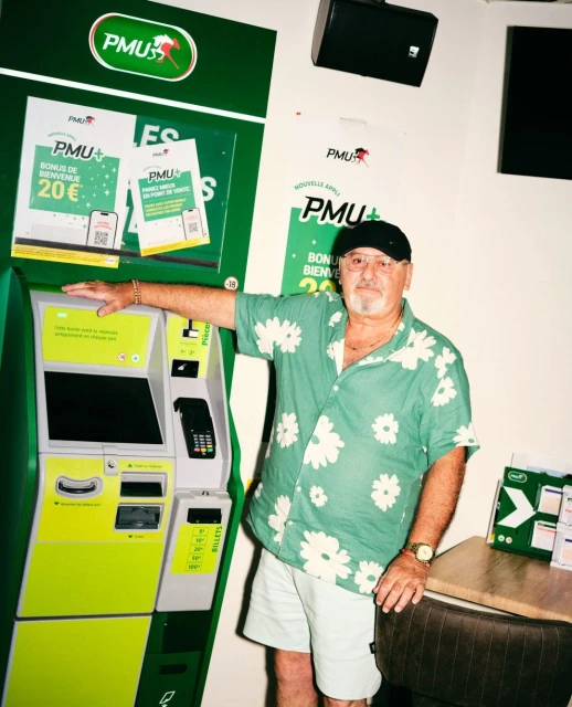 An adult man in a floral shirt stands beside a green and yellow PMU betting machine inside, with advertisements visible on the machine.