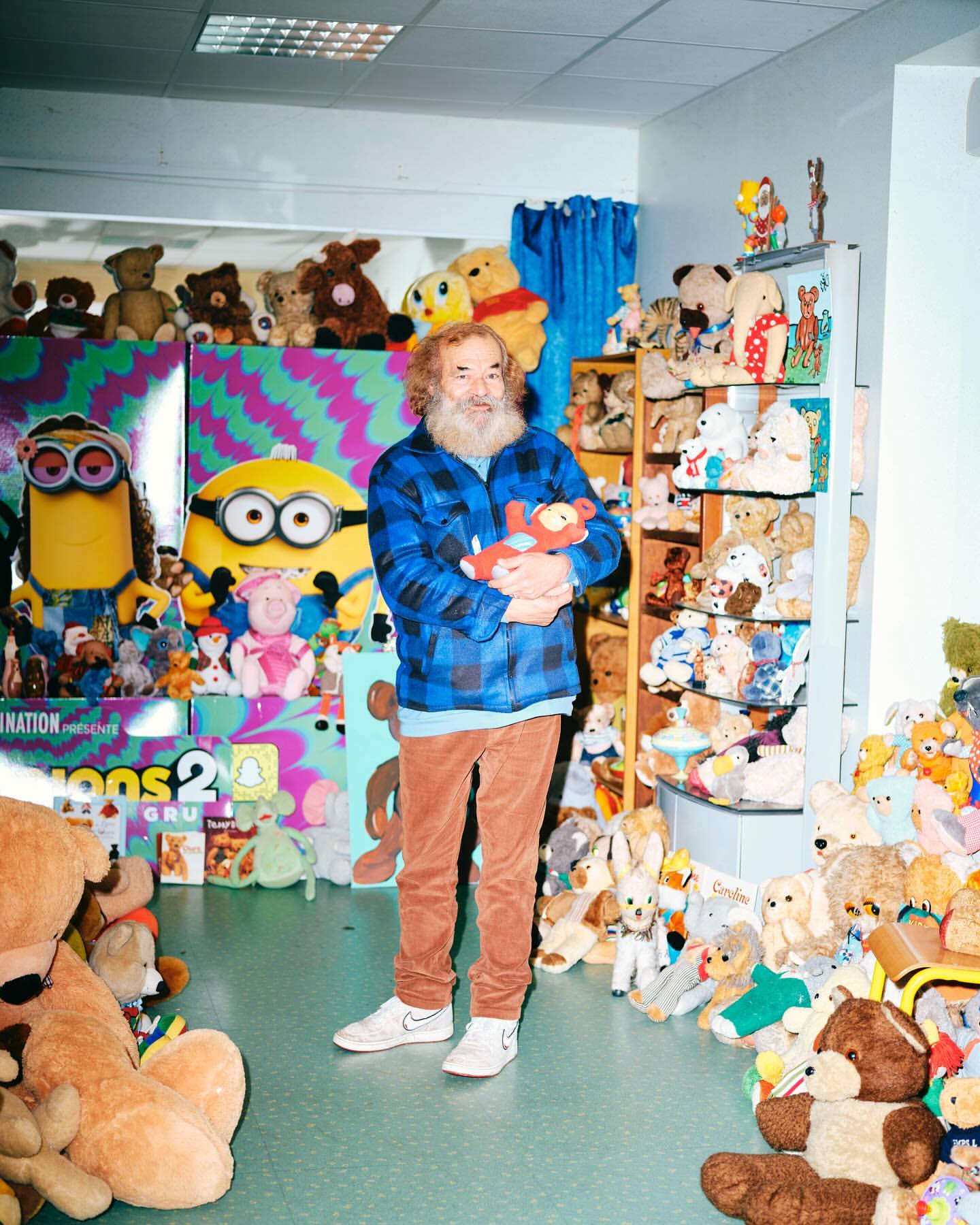 Adult man with a beard standing inside a room surrounded by teddy bears and character plush toys and holding a small bear.
