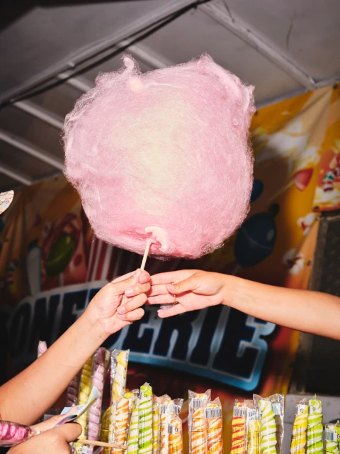 Two people exchanging a large, pink cotton candy at an outdoor fair. Colorful wrapped candies are displayed on the counter.
