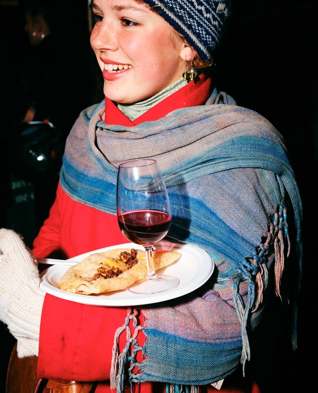 Smiling woman in winter hat and scarf holding plate with pastry and glass of red wine in dim lighting, creating a cozy atmosphere.