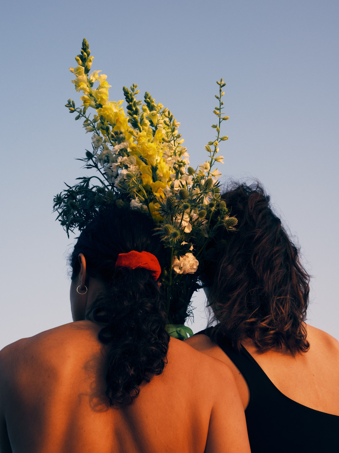 Two women with their backs to the camera, standing in an open space with a bouquet of yellow and white flowers against a clear sky.
