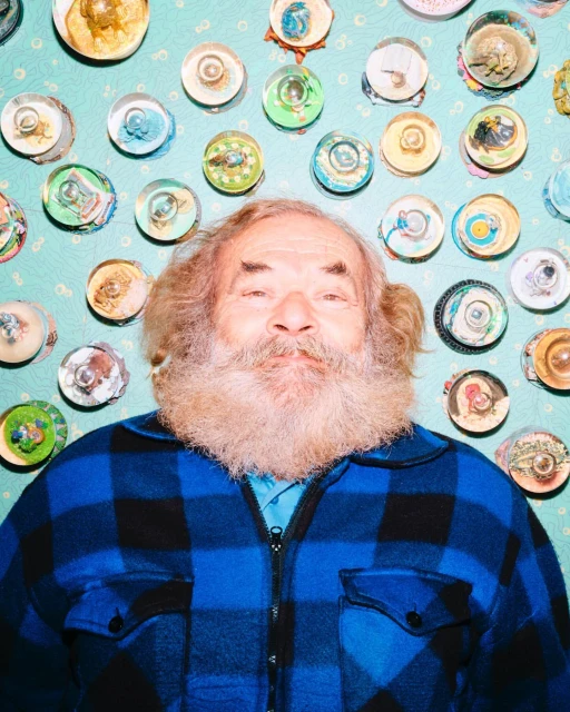 Close-up portrait of a bearded man in a blue plaid shirt, posing in front of a light blue wall decorated with small, colorful circular artworks.