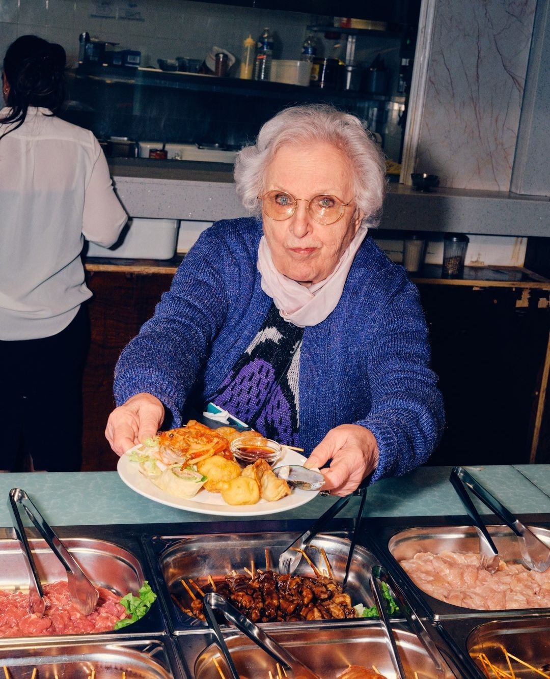 Senior woman in a blue sweater is holding a plate of food at a food buffet with tongs, metal trays, and various foods in the background.