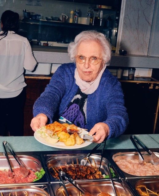 Senior woman in a blue sweater is holding a plate of food at a food buffet with tongs, metal trays, and various foods in the background.