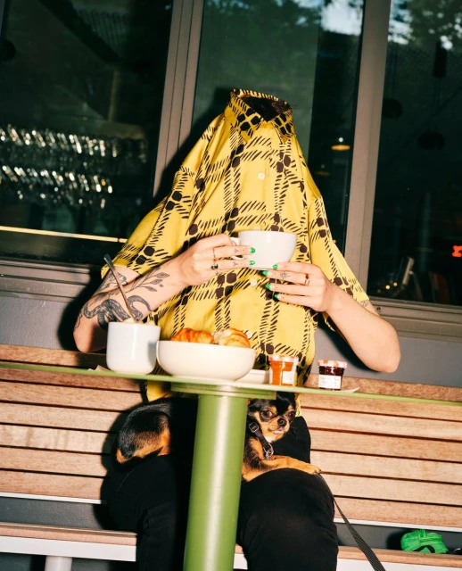 Person covered by a yellow pattern shirt holding a coffee cup at an outdoor cafe table with a small dog on the lap.