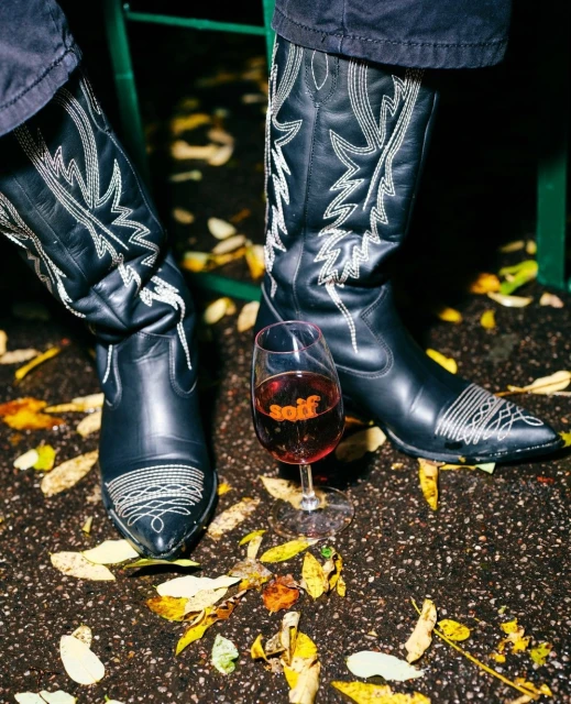 Close-up of black leather cowboy boots with white stitching, a wine glass with red wine, on a pavement strewn with autumn leaves.
