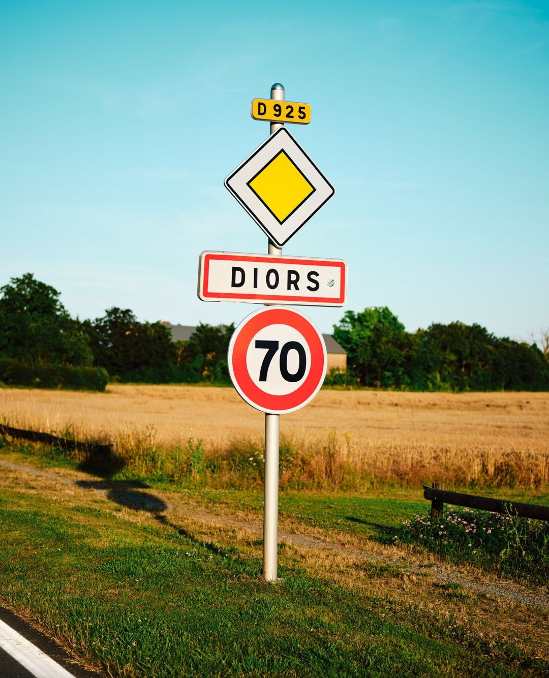 Roadside view featuring several stacked traffic signs indicating directions and speed limit against a clear blue sky and open field.