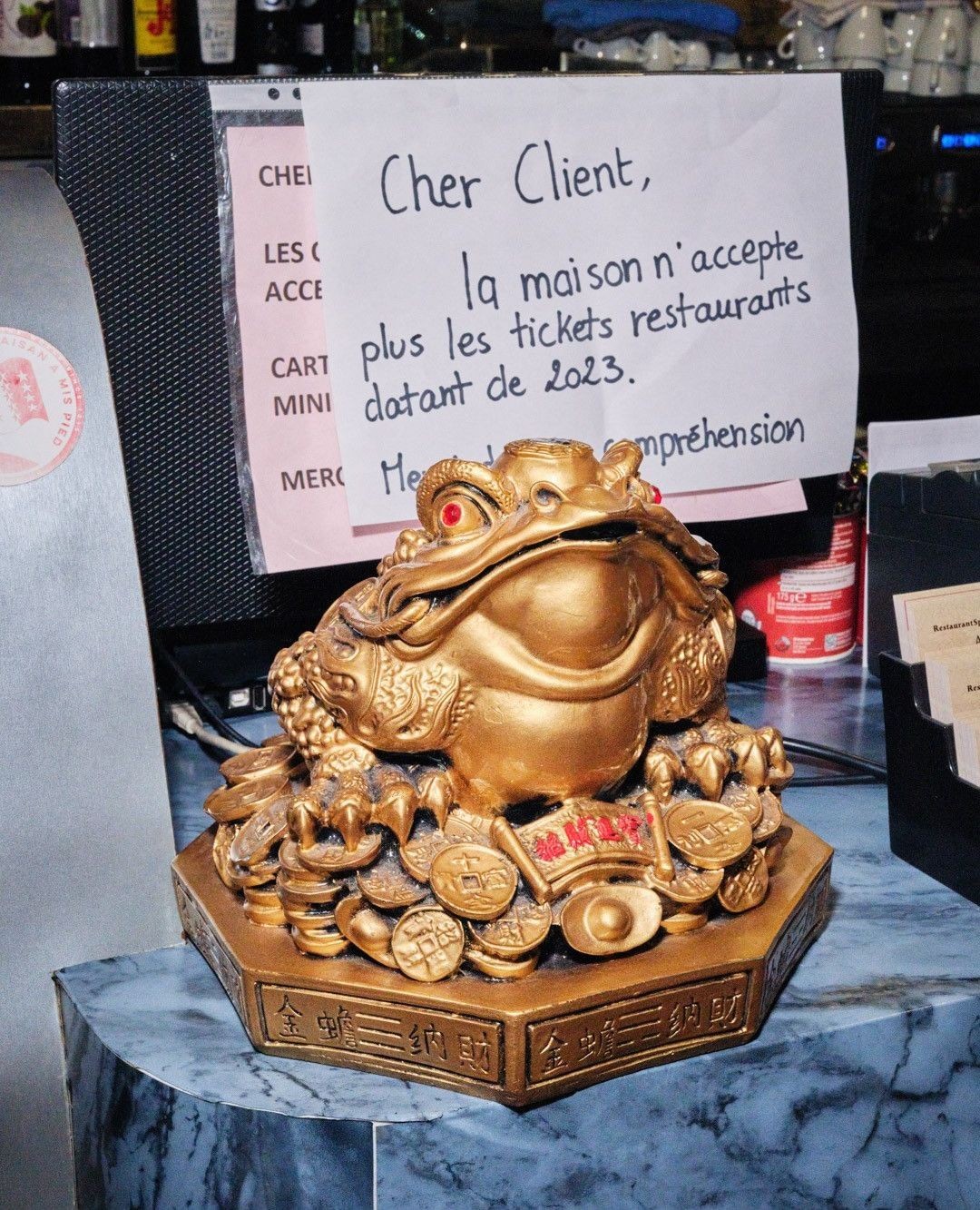 Golden money frog statue on a blue marble surface next to a sign in French, in an indoor setting with neutral lighting.