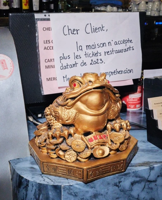 Golden money frog statue on a blue marble surface next to a sign in French, in an indoor setting with neutral lighting.